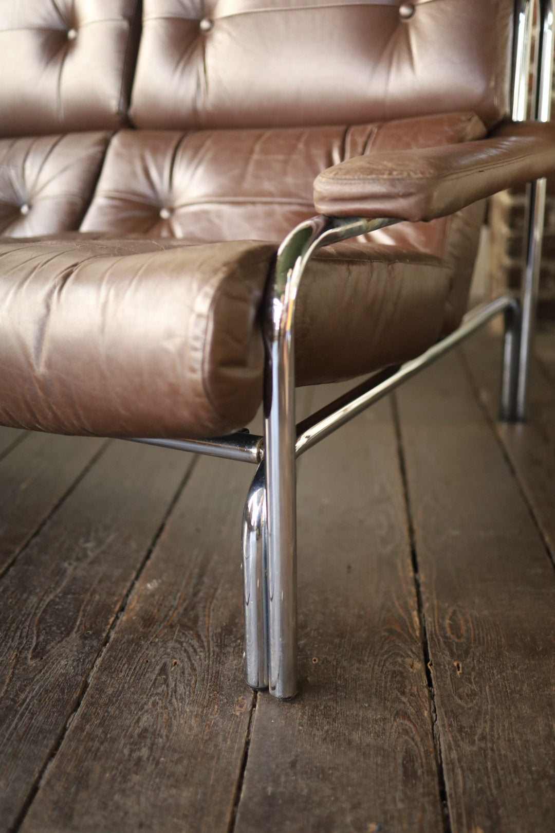 Close-up of the right armrest and seat of a vintage Pieff Alpha sofa, showing brown leather upholstery with button tufting and the shiny chrome leg on a wooden floor.