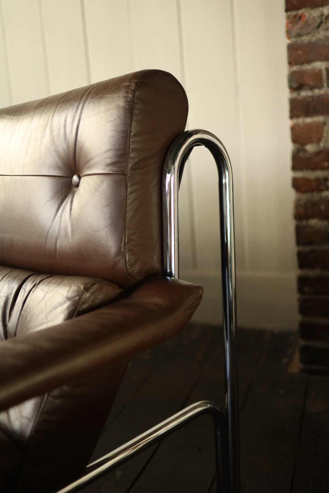 Detail of the upper corner of a Pieff Alpha sofa, showing brown leather button-tufted backrest and the curved chrome frame against a cream panelled wall and exposed brick.