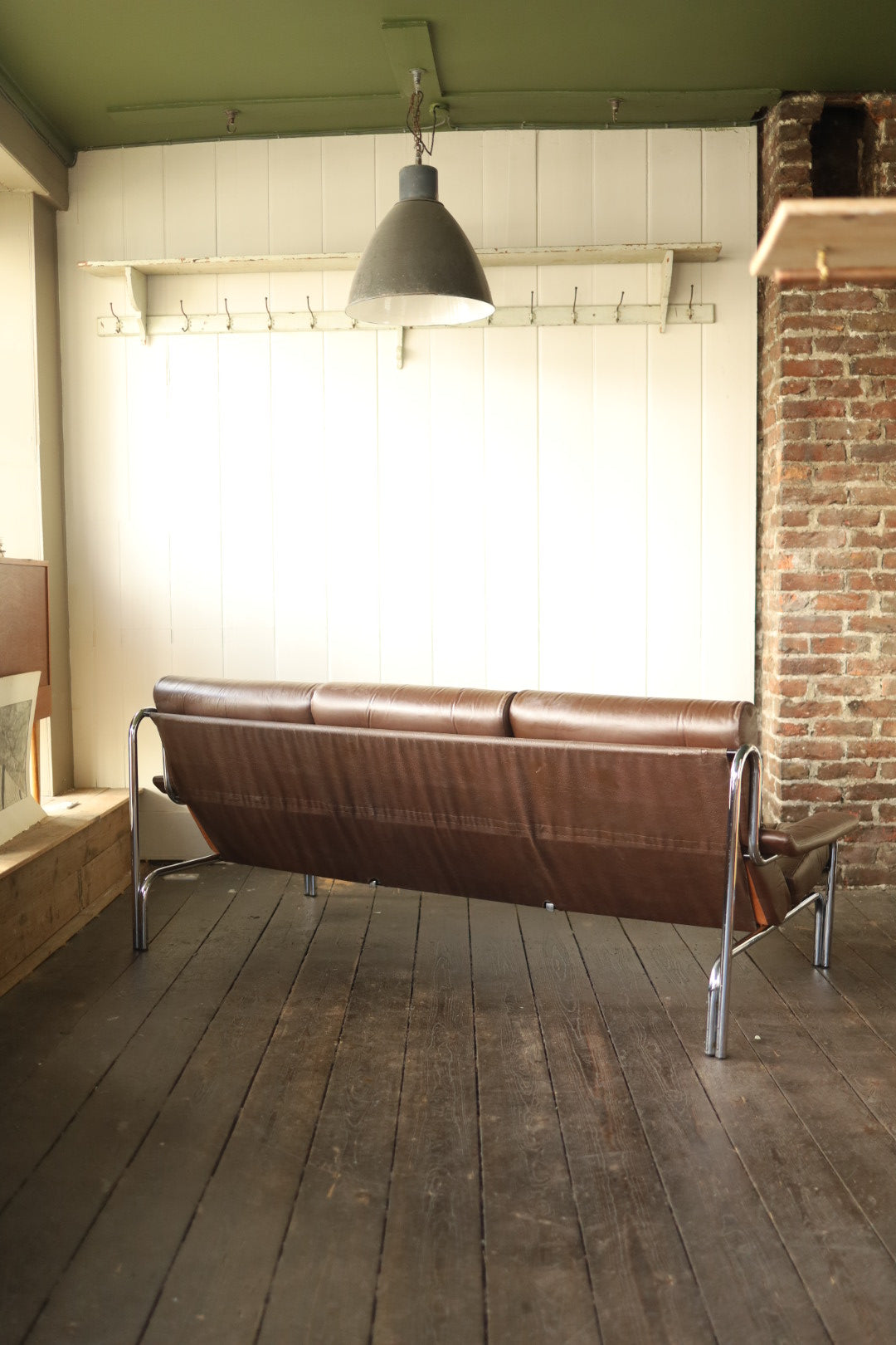 Rear view of a vintage Pieff Alpha three-seater sofa in brown leather with a chrome frame, placed in a light-filled room with wood flooring, white panelled walls, and a brick column.
