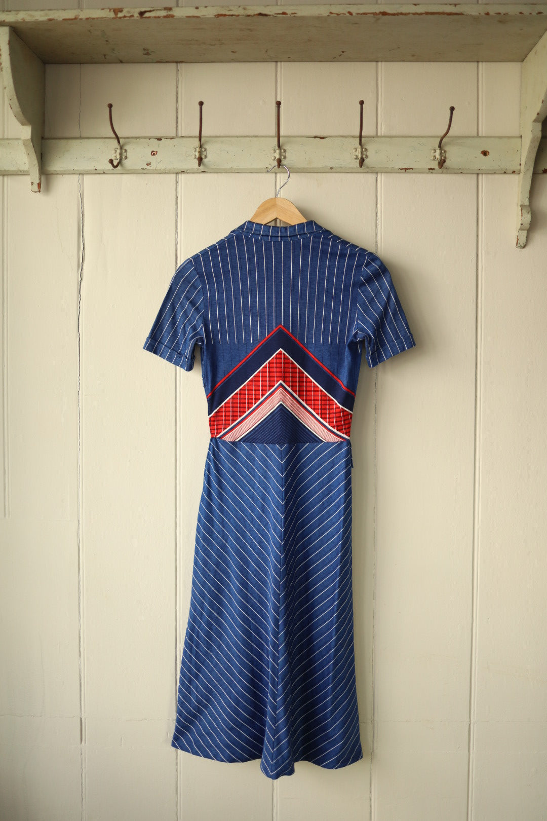 Back view of a vintage Betty Barclay 1970s short-sleeved dress, hanging on a wooden hanger against a painted wooden panel wall. The dress features vertical and diagonal white pinstripes on blue, with bold red, navy, and checked chevron panels across the waist for a striking graphic effect.