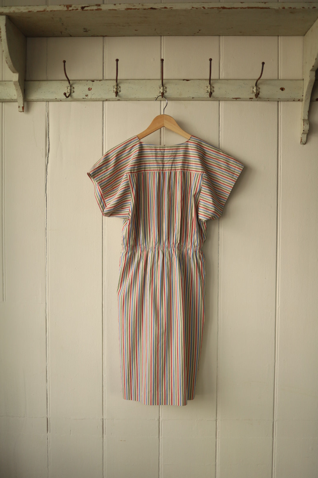 Back view of a handmade vintage dress with raglan sleeves, hanging on a wooden hanger against a painted wooden panel wall. The dress features a gathered elasticated waist and vertical multicoloured stripes on a light background, with a softly structured yoke across the shoulders.