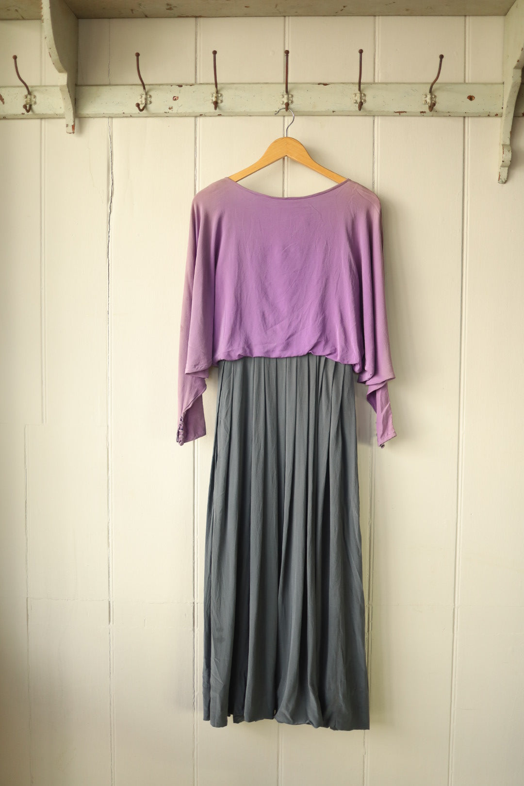 Front view of a John Bates silk dress with a soft heather top and pleated grey skirt, featuring wide sleeves, displayed on a wooden hanger against a cream panelled wall.