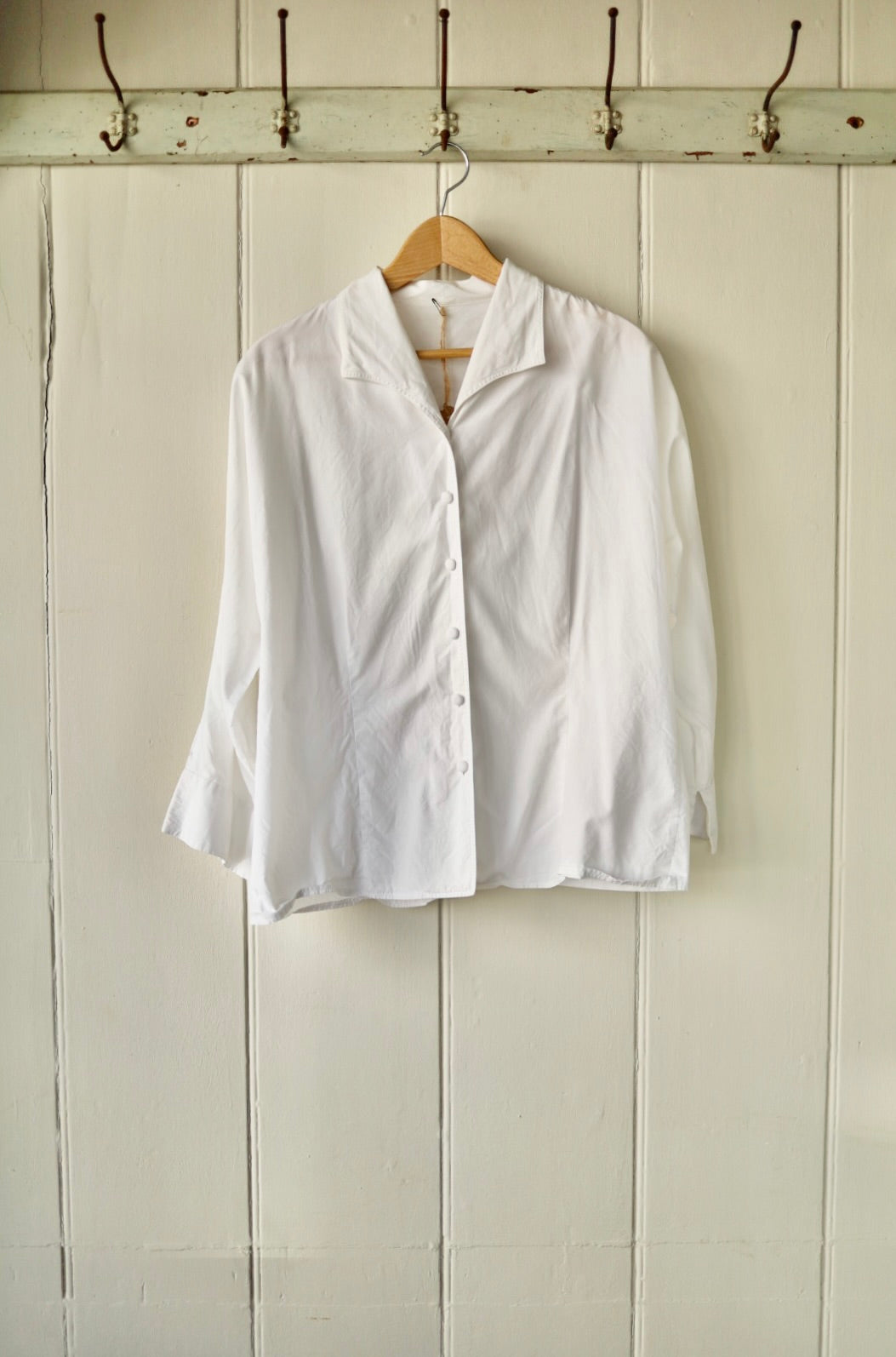 Vintage white blouse with shawl collar and fabric-covered buttons, displayed on wooden hanger against a painted tongue-and-groove wall.