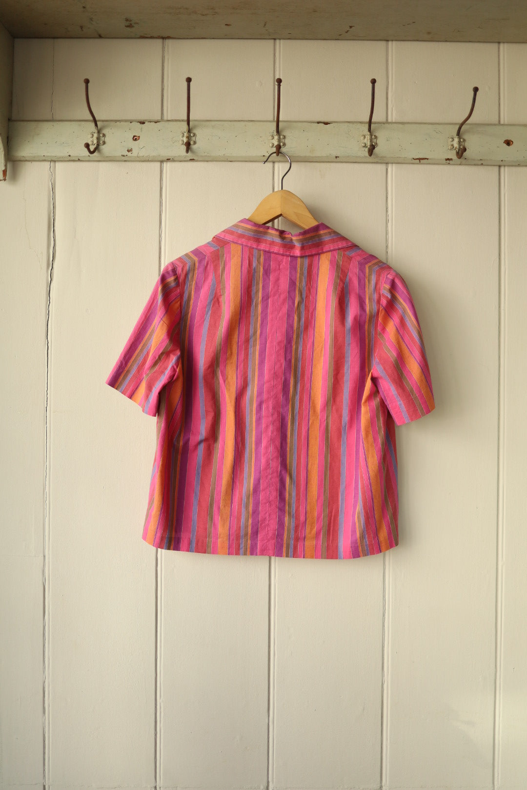 Back view of the same candy stripe shirt, showing the continuation of the vivid vertical stripe pattern and the structured back seam, also hung on a wooden hanger against a white panelled wall.