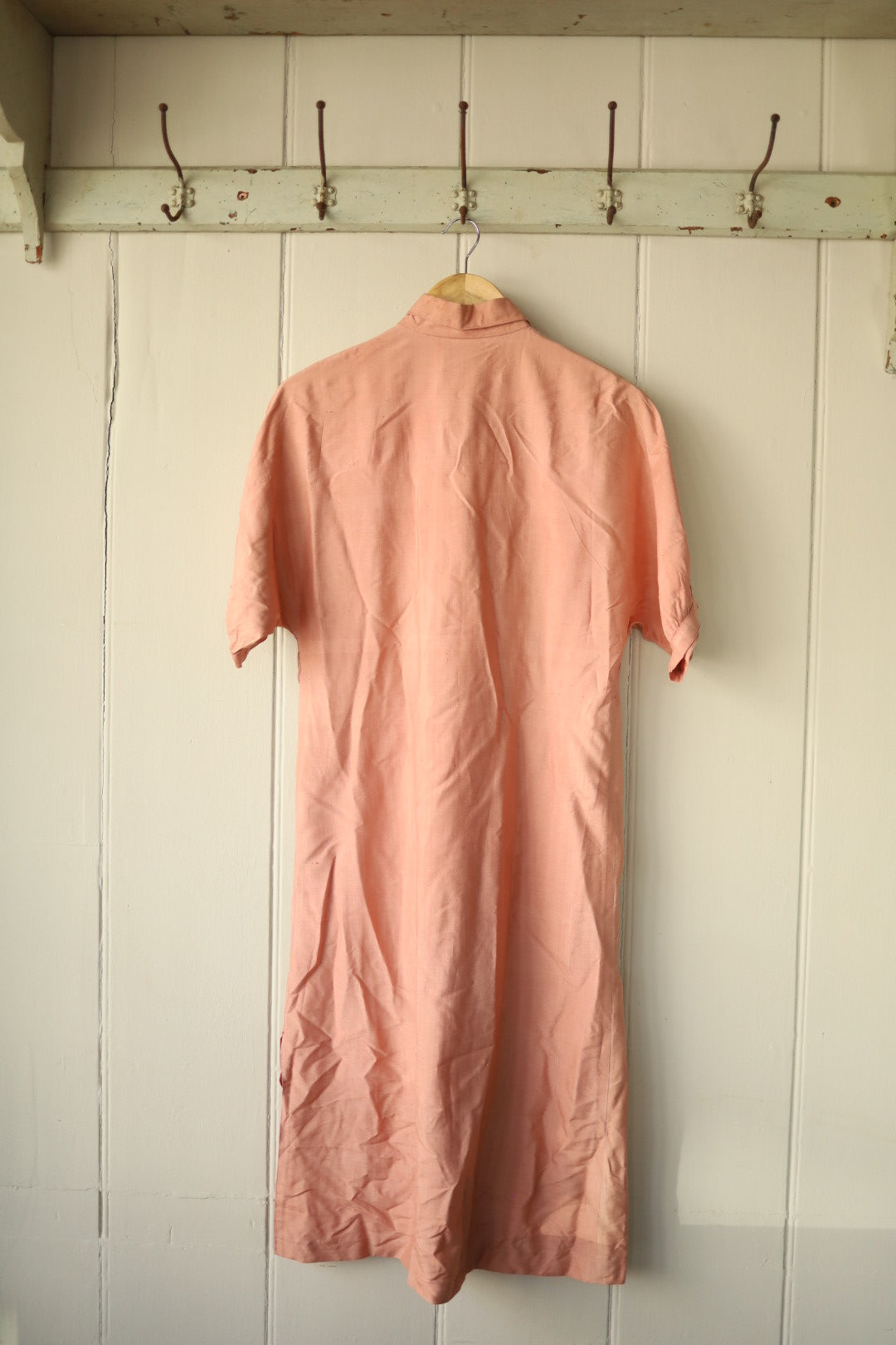 Back of the vintage nude silk dress showing its simple clean lines and short sleeves, displayed on a wooden hanger against a white panelled backdrop.