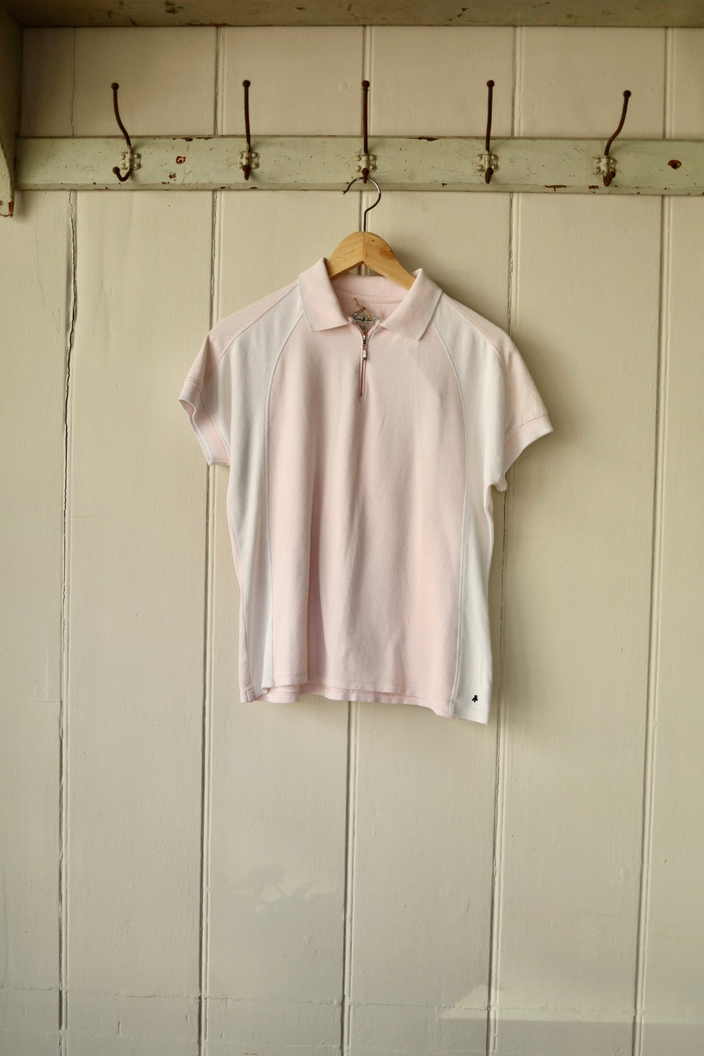Vintage blush pink and white cotton tennis top with short raglan sleeves and a half-zip neckline, displayed on a wooden hanger against a cream panelled wall.