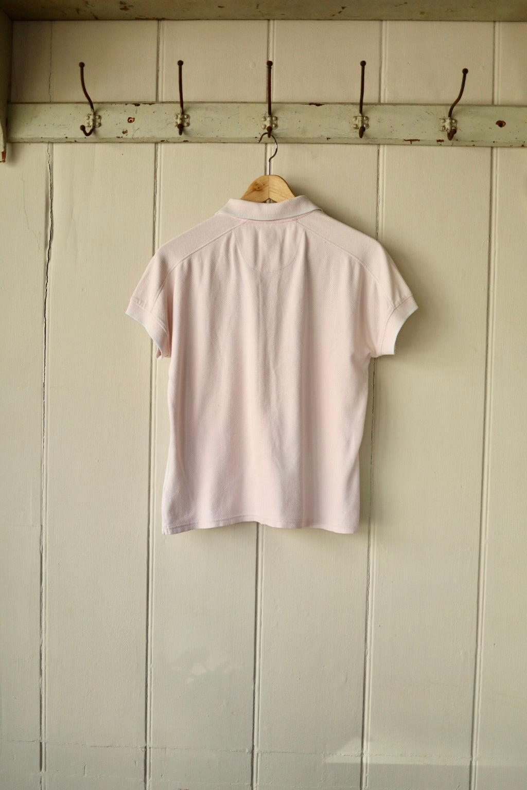 Back of the vintage tennis top showing plain blush pink cotton with raglan sleeve construction, hanging neatly on a wooden hanger against a cream panelled wall.
