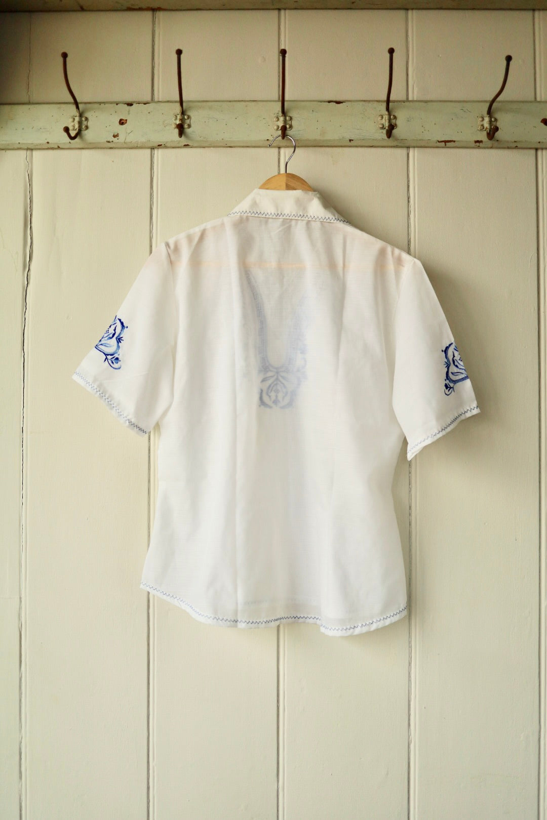 Back view of the vintage white embroidered shirt, showing the semi-sheer cotton fabric and subtle reverse of the blue front embroidery, displayed on a wooden hanger against a cream panelled wall.