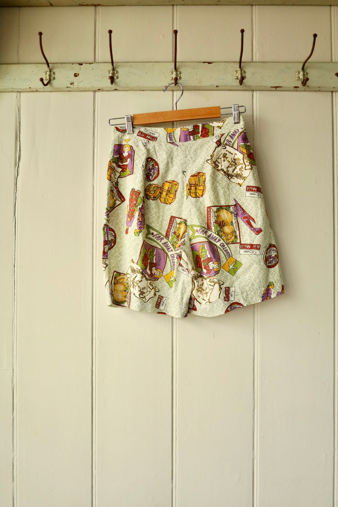 Back view of the same vintage printed shorts, showing an all-over novelty travel print featuring maps, postcards, and luggage motifs, hung on a wooden hanger against a cream wall with iron hooks.