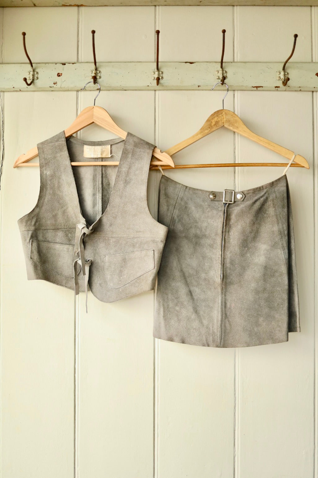 Back view of a vintage 1970s grey suede two-piece set, featuring a cropped sleeveless vest and A-line mini skirt, both hanging on wooden hangers against a painted wooden wall with hooks.