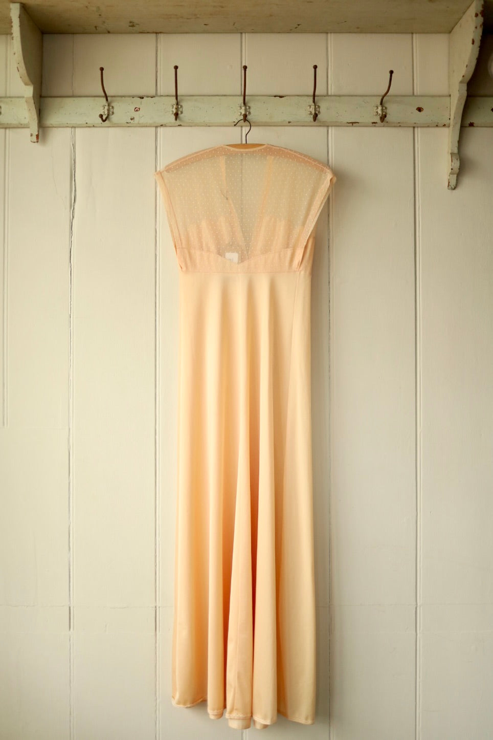 Back view of a vintage peach nightdress showing sheer mesh panel across the shoulders and back, displayed on a wooden hanger against a pale wooden wall