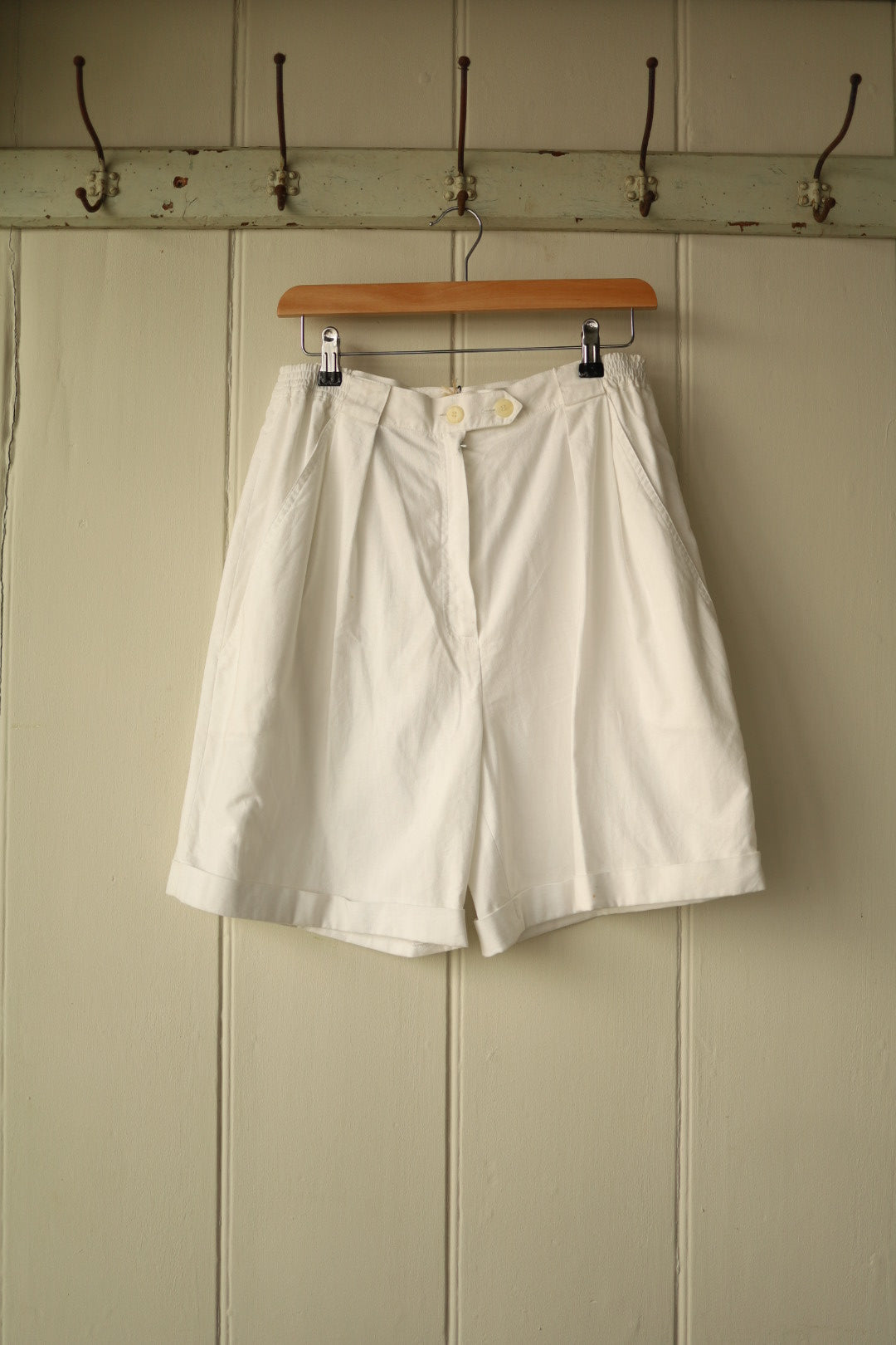 Front view of cream high-waisted vintage shorts with pleats, double button fastening, and turned-up hem, hanging on wooden hanger against a neutral panelled wall.