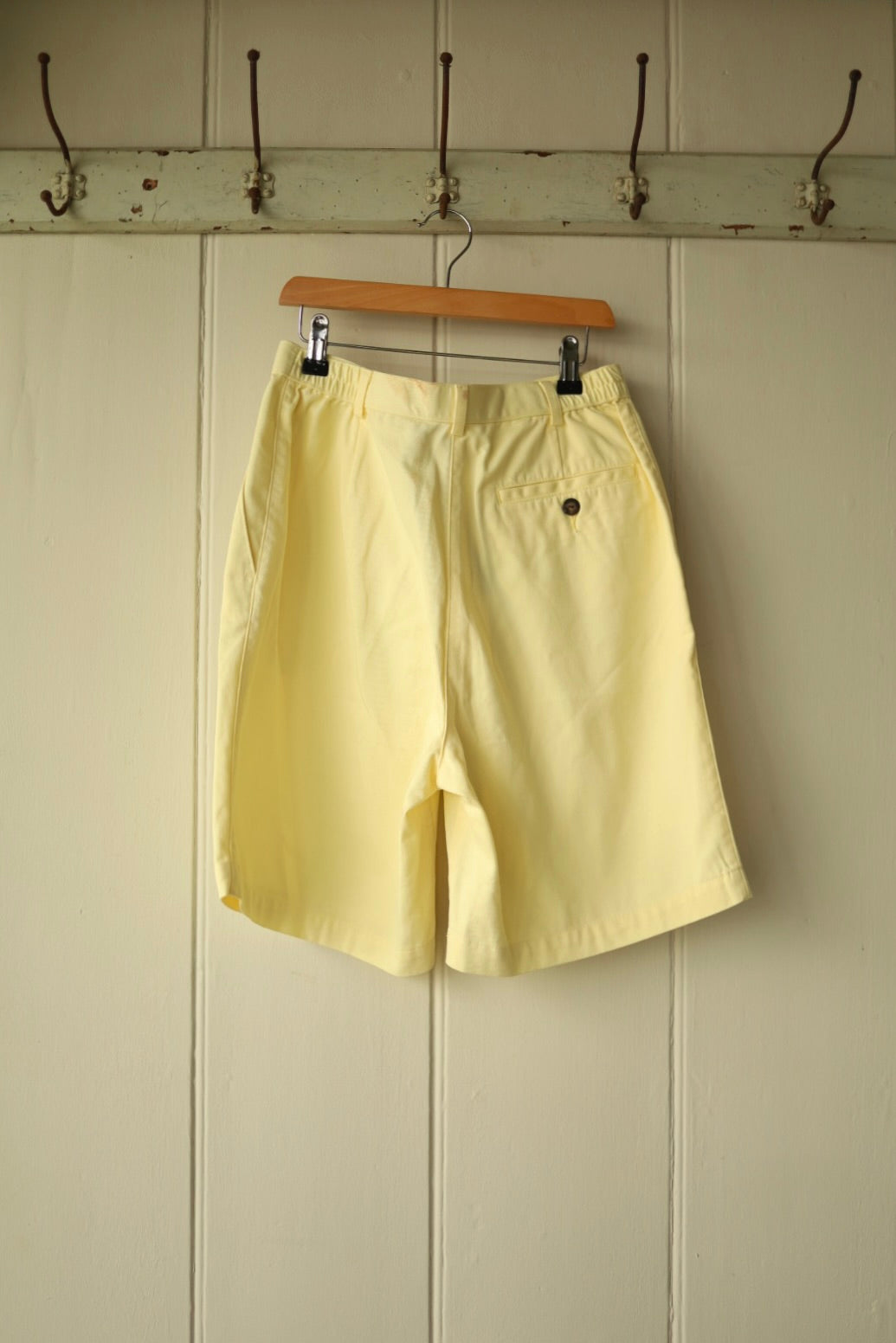 Back view of vintage lemon yellow pleated shorts showing elasticated side waistband and single back pocket with button, displayed on a wooden hanger against a cream panelled wall.