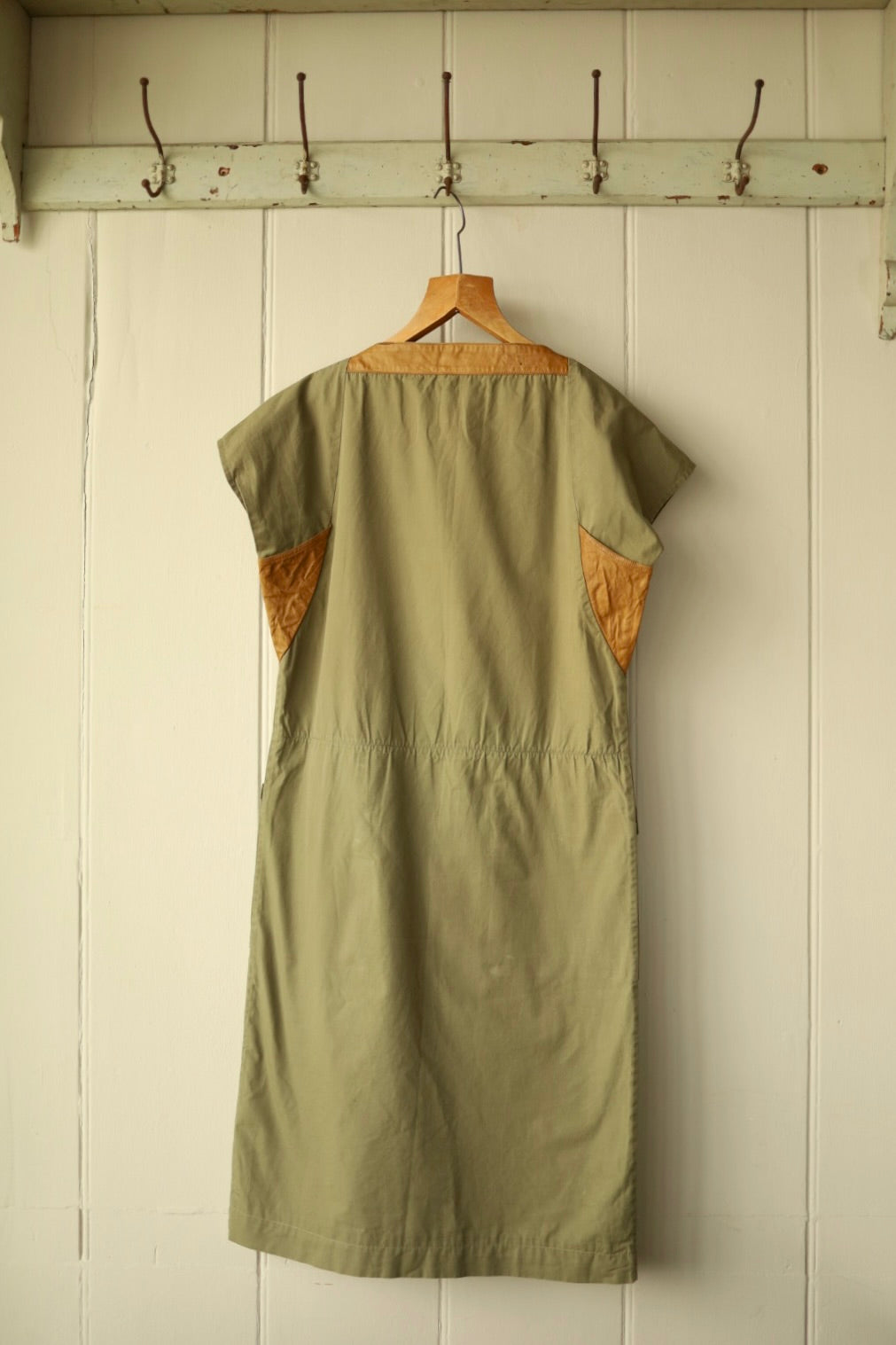Back view of the khaki dress with tan yoke and underarm inserts, shown against a cream painted wooden wall.