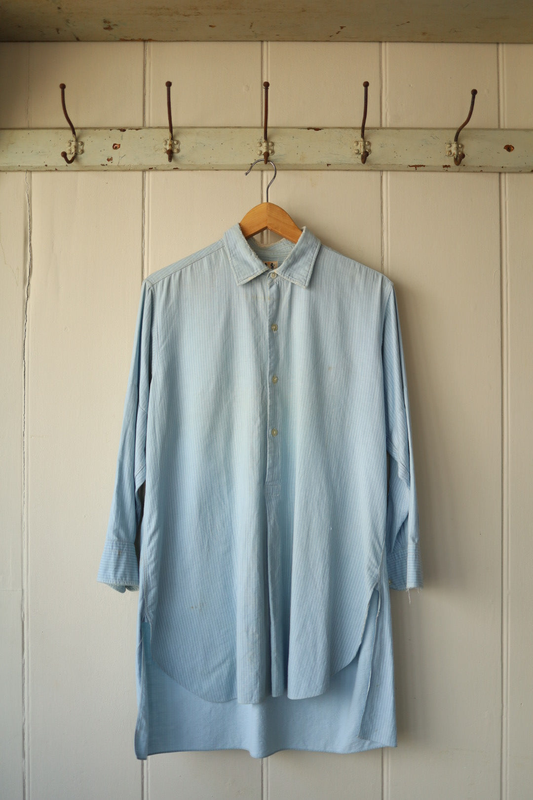 Front view of oversized pale blue striped shirt hanging against a vintage cream panelled wall with hooks above.