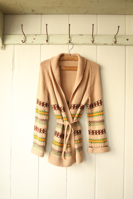 A vintage knitted cardigan in warm beige with colourful Fair Isle-style horizontal patterns in red, green, yellow, and brown. The cardigan features a shawl collar, long sleeves, and a matching knitted belt tied at the waist. Displayed on a vintage wooden hanger against a cream tongue and groove wall with old metal coat hooks above.