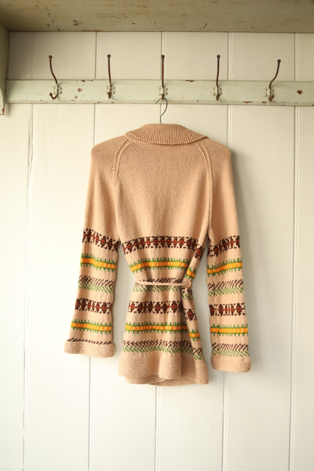 Back view of the same vintage knitted cardigan, showing the colourful Fair Isle-style detailing wrapping around the waist and arms. The knitted belt is tied at the back, and the garment is hung on a vintage wooden hanger against a pale wood-panelled wall.