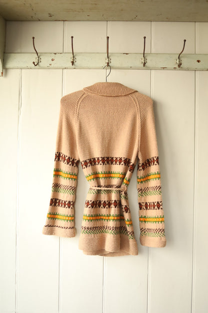 Back view of the same vintage knitted cardigan, showing the colourful Fair Isle-style detailing wrapping around the waist and arms. The knitted belt is tied at the back, and the garment is hung on a vintage wooden hanger against a pale wood-panelled wall.