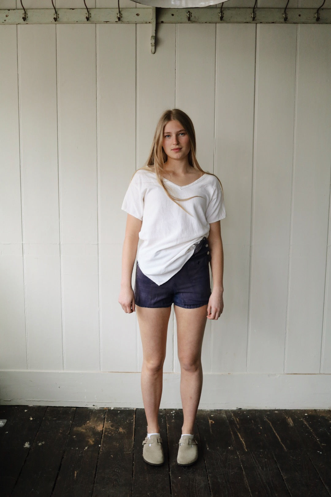 Full body shot of a model wearing vintage 1930s blue workwear shorts with a tucked white textured tee and Birkenstock-style clogs, standing against a painted wood-panel wall.