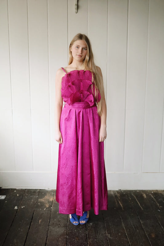 Woman wearing a vibrant fuchsia pink vintage evening dress, standing indoors against a painted wooden panel wall. The dress features slim shoulder straps, dramatic pleated fan detailing across the bodice, and a full-length skirt with a moiré pattern. She pairs it with cobalt blue heeled sandals on a dark wooden floor.