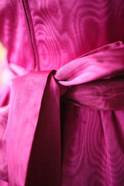 Close-up detail of a fuchsia pink vintage dress showing the moiré-patterned fabric and a neatly tied waistband bow. The textured swirl of the fabric is clearly visible, highlighting the rich colour and sheen