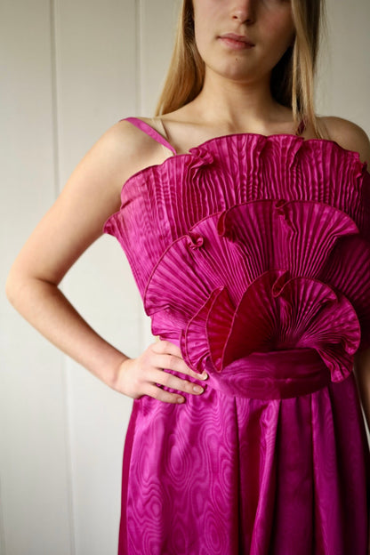 Close-up of a woman wearing a fuchsia pink vintage evening dress, focusing on the intricately pleated fan detailing across the bodice. The dress features a wide waistband and moiré-patterned fabric, with the model's hand resting on her hip.
