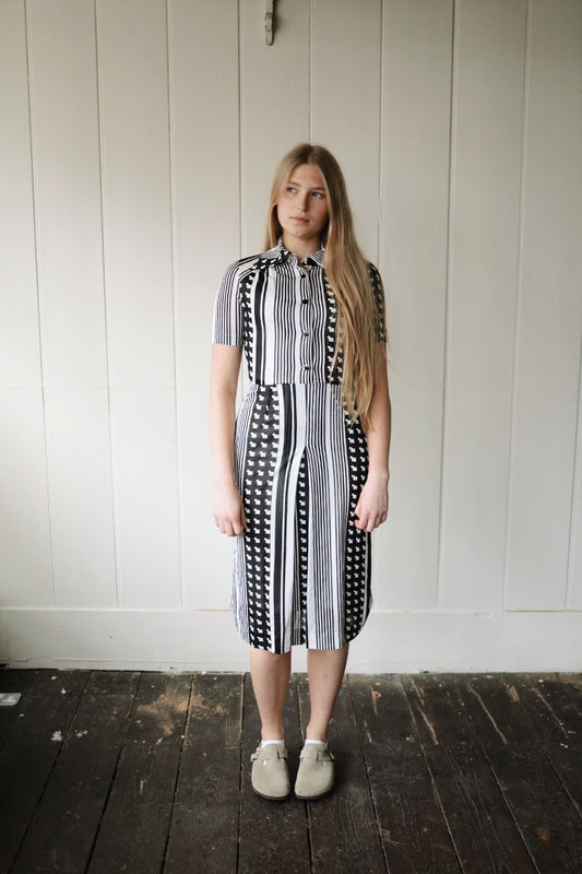 Woman wearing a vintage monochrome dress standing indoors against a painted wooden panel wall. The dress features vertical black and white stripes with a bold graphic motif, short sleeves, a fitted bodice, and flared skirt. She pairs it with neutral slip-on shoes on a dark wooden floor.