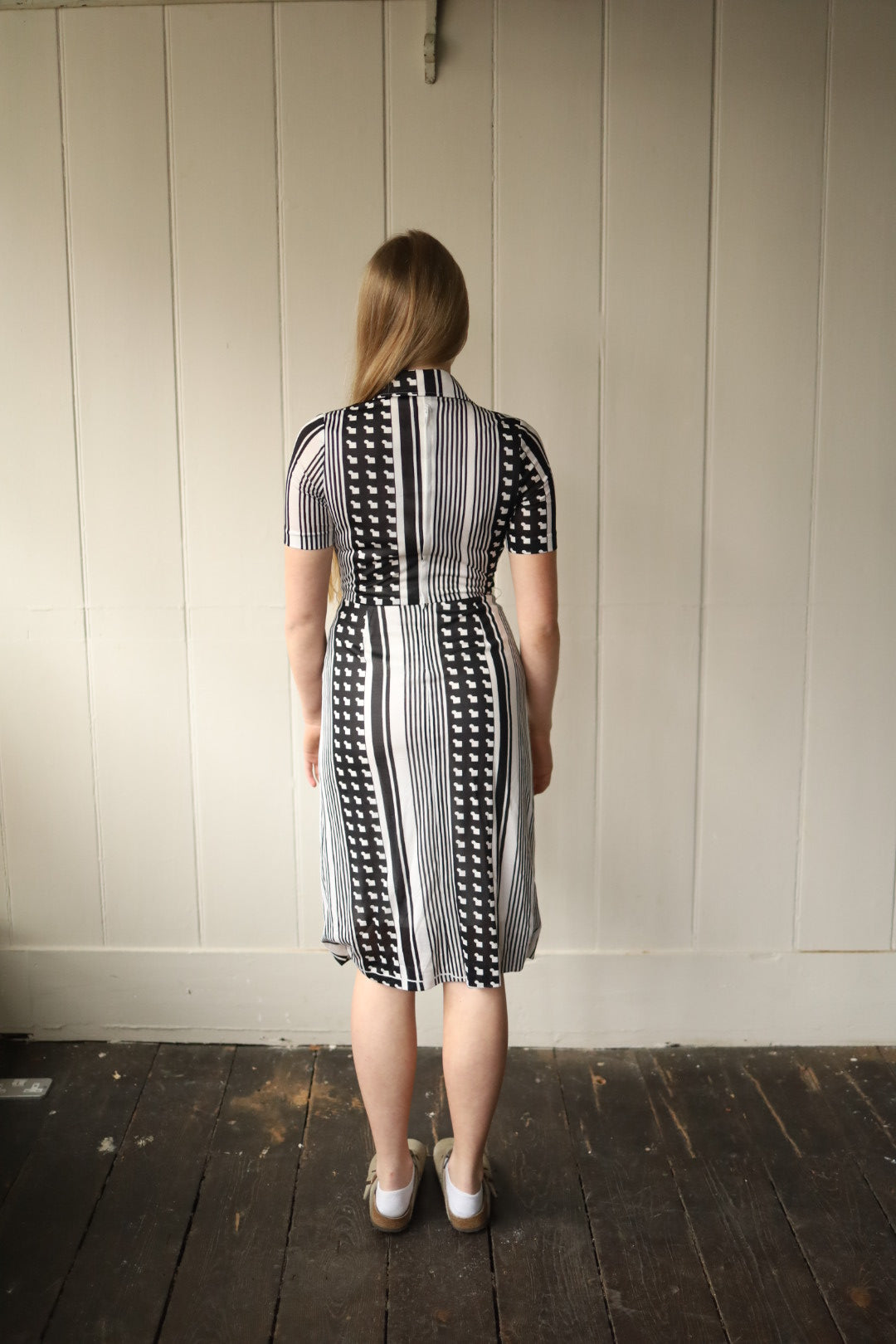 Back view of a woman wearing a vintage monochrome dress, standing indoors on a dark wooden floor against a painted panel wall. The dress features vertical black and white stripes combined with a bold geometric motif, fitted through the bodice with a gently flared skirt and short sleeves.