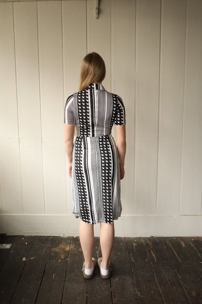 Back view of a woman wearing a vintage monochrome dress, standing indoors on a dark wooden floor against a painted panel wall. The dress features vertical black and white stripes combined with a bold geometric motif, fitted through the bodice with a gently flared skirt and short sleeves.