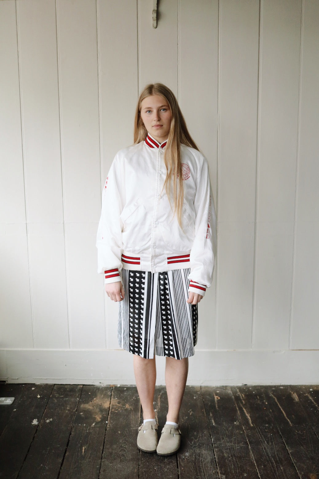 Front view of a young woman standing against a white wooden wall, wearing a white bomber jacket with red striped collar, cuffs, and hem, styled with a monochrome geometric pleated skirt and beige slip-on shoes.