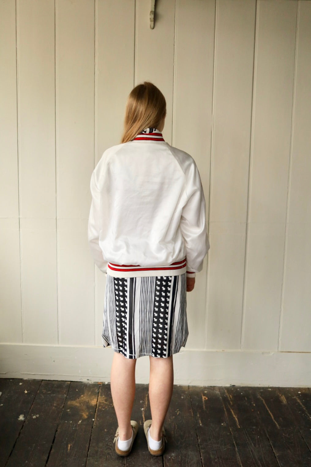 Rear view of the same outfit, showing the back of the white bomber jacket and the skirt, with the model’s long blonde hair falling straight down.