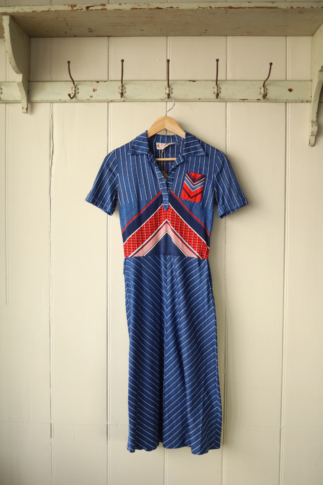 Vintage Betty Barclay 1970s short-sleeved dress hanging on a wooden hanger against a painted wooden panel wall. The dress features a pointed collar, buttoned neckline, and bold geometric panel design in red, white, and blue with vertical and diagonal stripes.