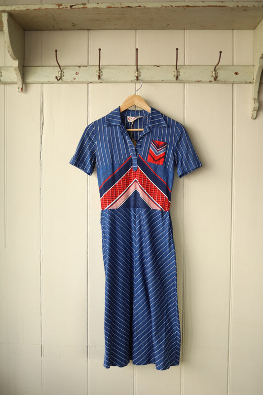 Vintage Betty Barclay 1970s short-sleeved dress hanging on a wooden hanger against a painted wooden panel wall. The dress features a pointed collar, buttoned neckline, and bold geometric panel design in red, white, and blue with vertical and diagonal stripes.