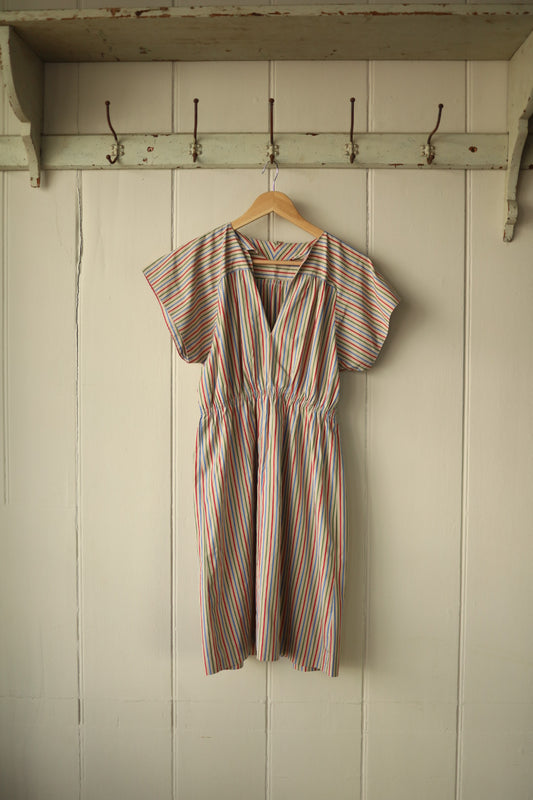 Handmade vintage dress with raglan sleeves, hanging on a wooden hanger against a painted wooden panel wall. The dress features a deep V-neckline, elasticated waist, and multi-coloured vertical stripes on a light cream background.