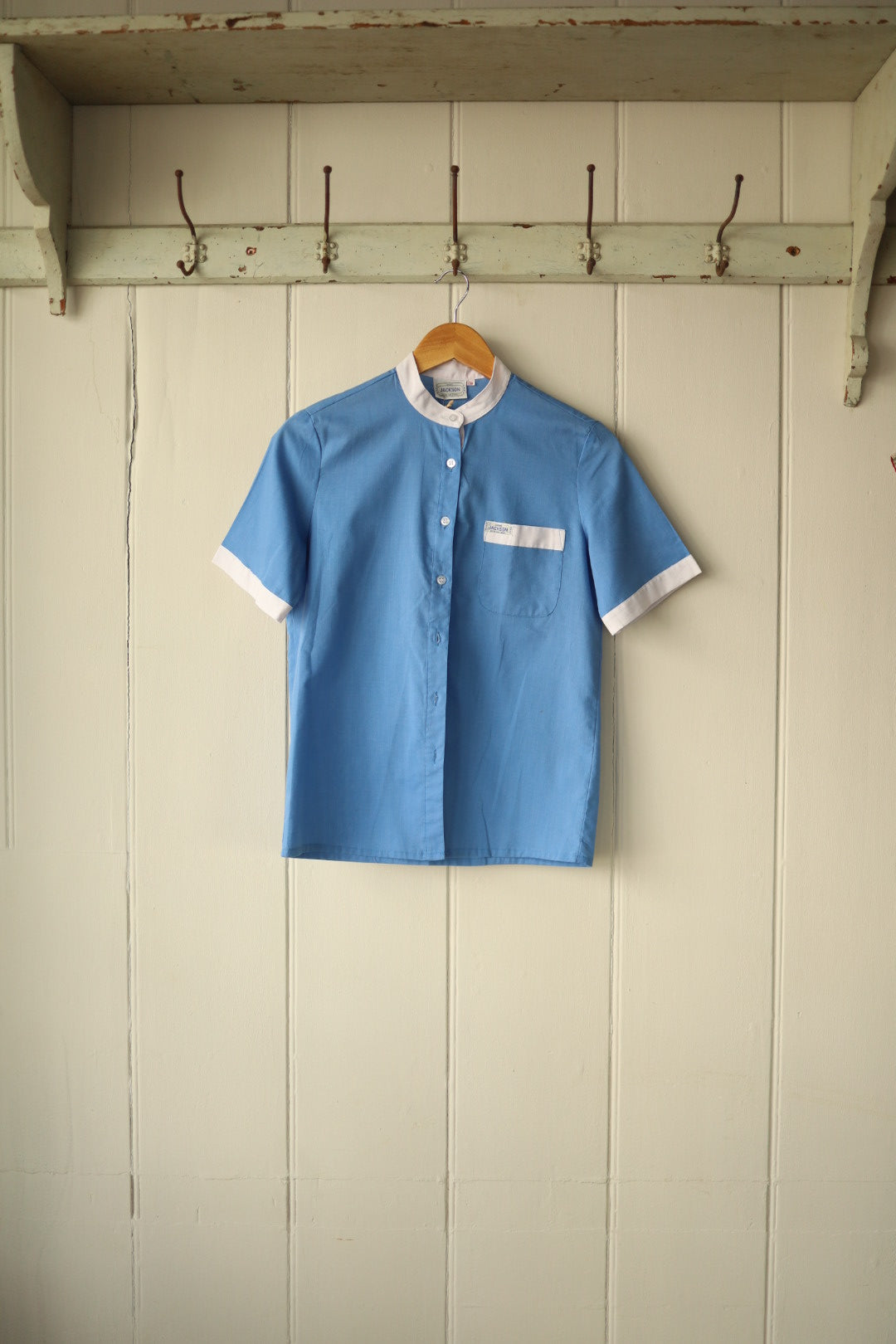 Vintage Jackson short-sleeved shirt hanging on a wooden hanger against a painted wooden panel wall. The shirt is sky blue with white contrast trim at the cuffs, collar, and pocket edge. Features a mandarin collar and front button fastening.