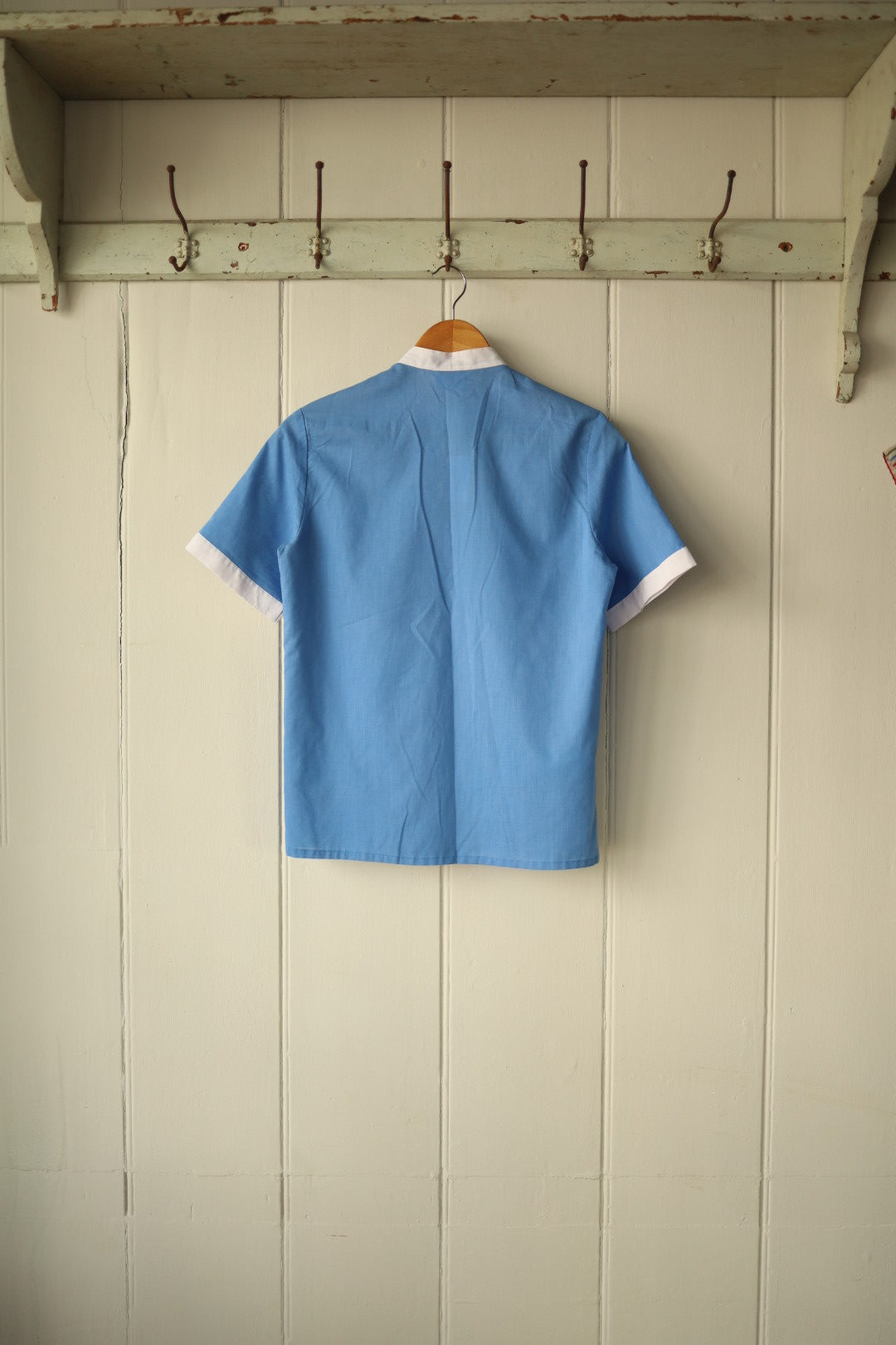 Back view of a vintage Jackson short-sleeved shirt, hanging on a wooden hanger against a painted wooden panel wall. The shirt is sky blue with white contrast trim at the sleeve cuffs and collar, featuring clean lines and a simple, structured cut.