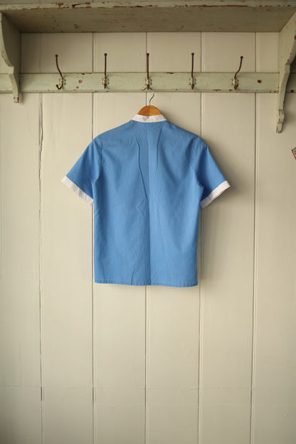 Back view of a vintage Jackson short-sleeved shirt, hanging on a wooden hanger against a painted wooden panel wall. The shirt is sky blue with white contrast trim at the sleeve cuffs and collar, featuring clean lines and a simple, structured cut.
