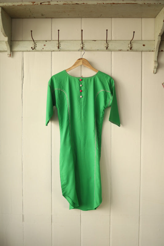Front view of a vintage leaf green dress hanging on a wooden hanger against a painted wooden panel wall. The dress features contrast stripe seam detailing along the shoulders and bodice, and a row of decorative buttons in red and white down the front neckline. It has short sleeves and a gently tailored silhouette.
