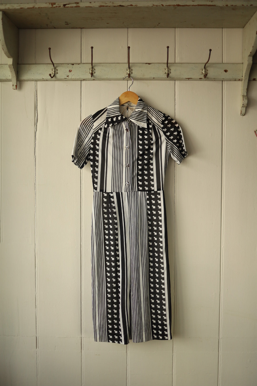 Front view of a vintage monochrome dress hanging on a wooden hanger against a painted wooden panel wall. The dress features a bold black and white pattern of vertical stripes and graphic motifs, with short sleeves, a fitted bodice, and a flared skirt. It has a wide pointed collar and front button fastening.