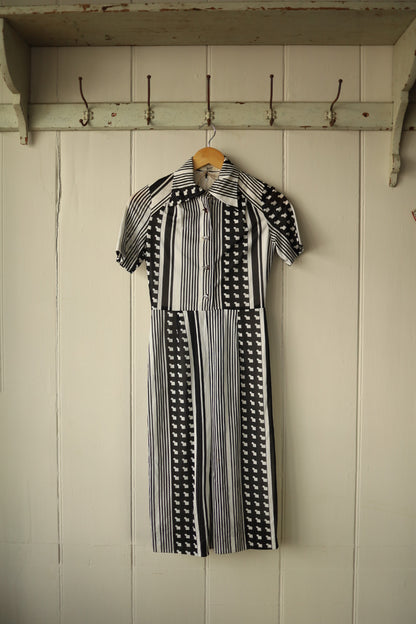 Front view of a vintage monochrome dress hanging on a wooden hanger against a painted wooden panel wall. The dress features a bold black and white pattern of vertical stripes and graphic motifs, with short sleeves, a fitted bodice, and a flared skirt. It has a wide pointed collar and front button fastening.
