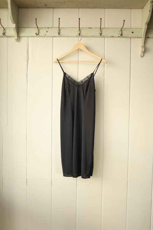 Black vintage slip dress with lace trim on neckline, hanging on wooden hanger against cream panelled wall.