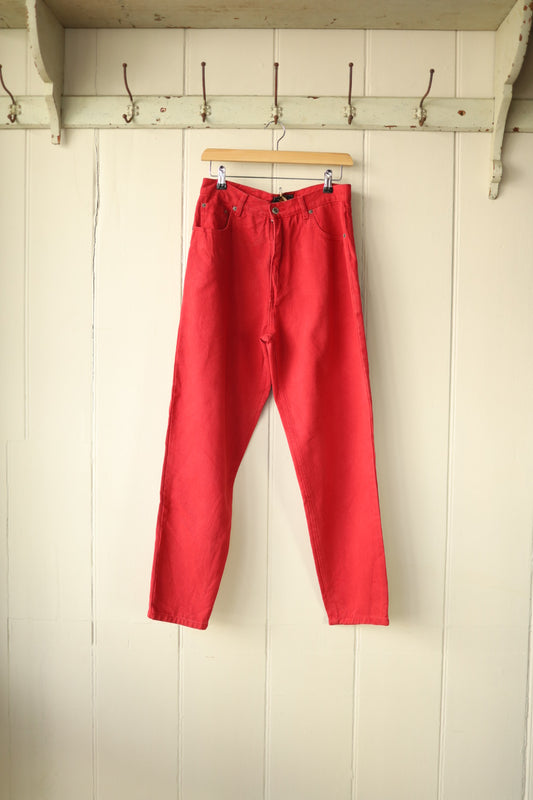 Front view of bright red Route 66 jeans hanging on a wooden hanger against a cream panelled wall.