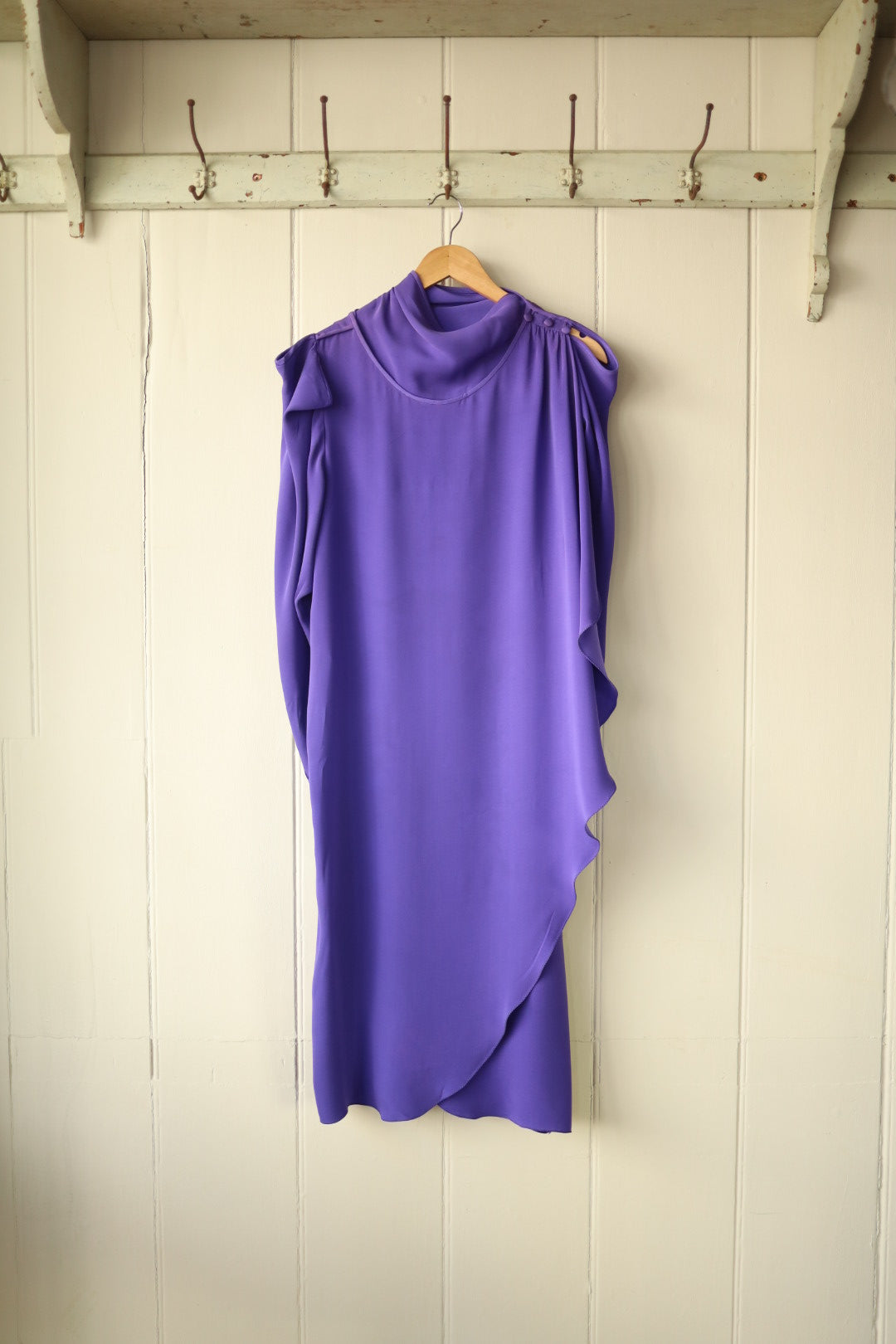 Purple asymmetrical draped dress with high neckline and cold shoulder detail, hanging on a wooden hanger against a cream panelled wall with vintage hooks.