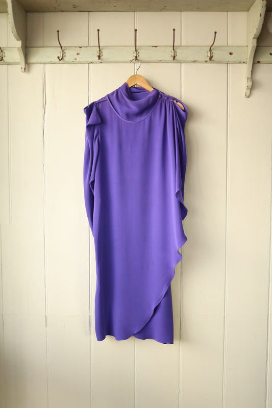 Purple asymmetrical draped dress with high neckline and cold shoulder detail, hanging on a wooden hanger against a cream panelled wall with vintage hooks.