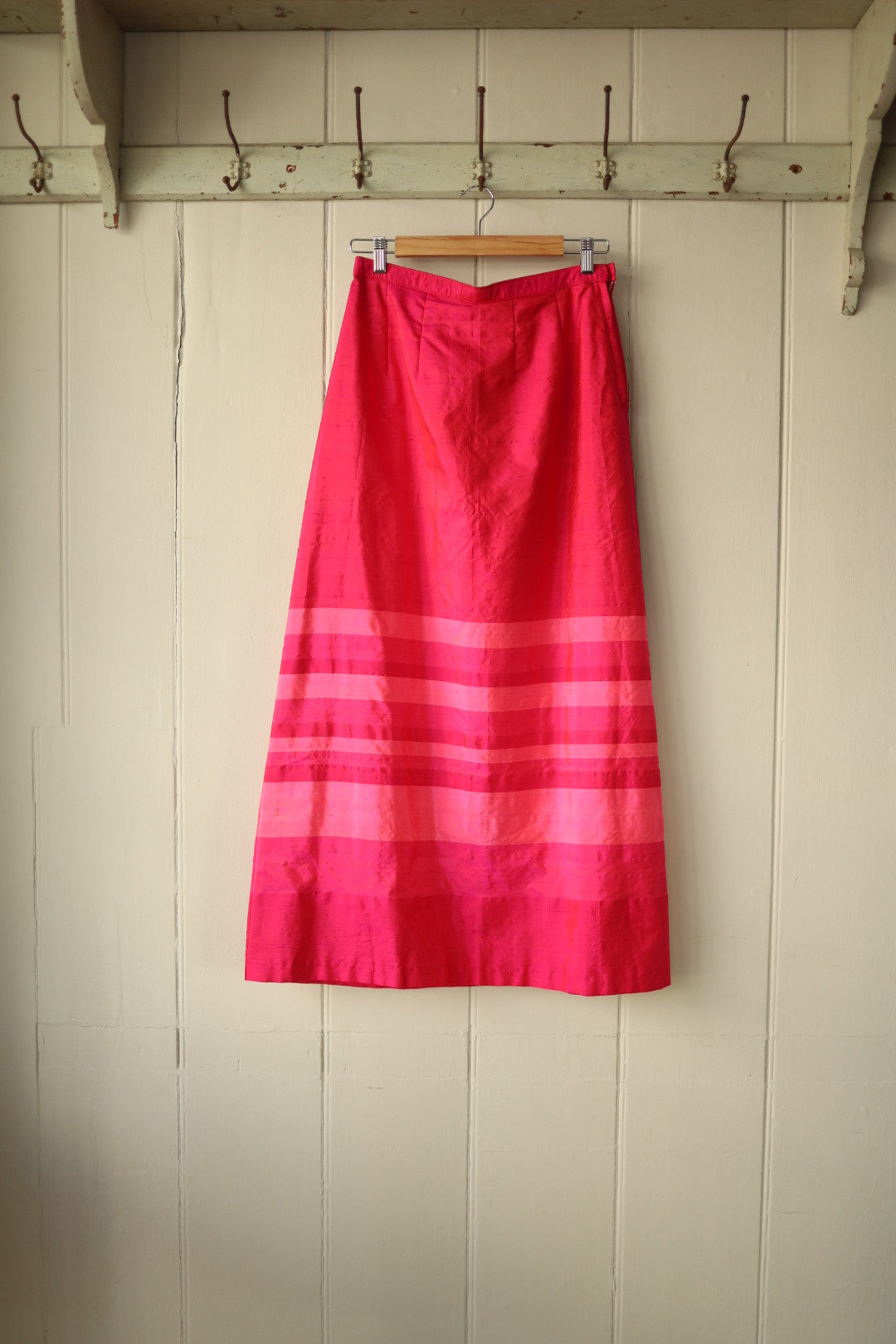 Fuschia silk maxi skirt with horizontal tonal stripes near the hem, hanging on a wooden hanger against a cream panelled wall.