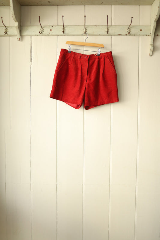 Front view of high-waisted red suede shorts with side pockets, hanging on a wooden hanger against a cream panelled wall.