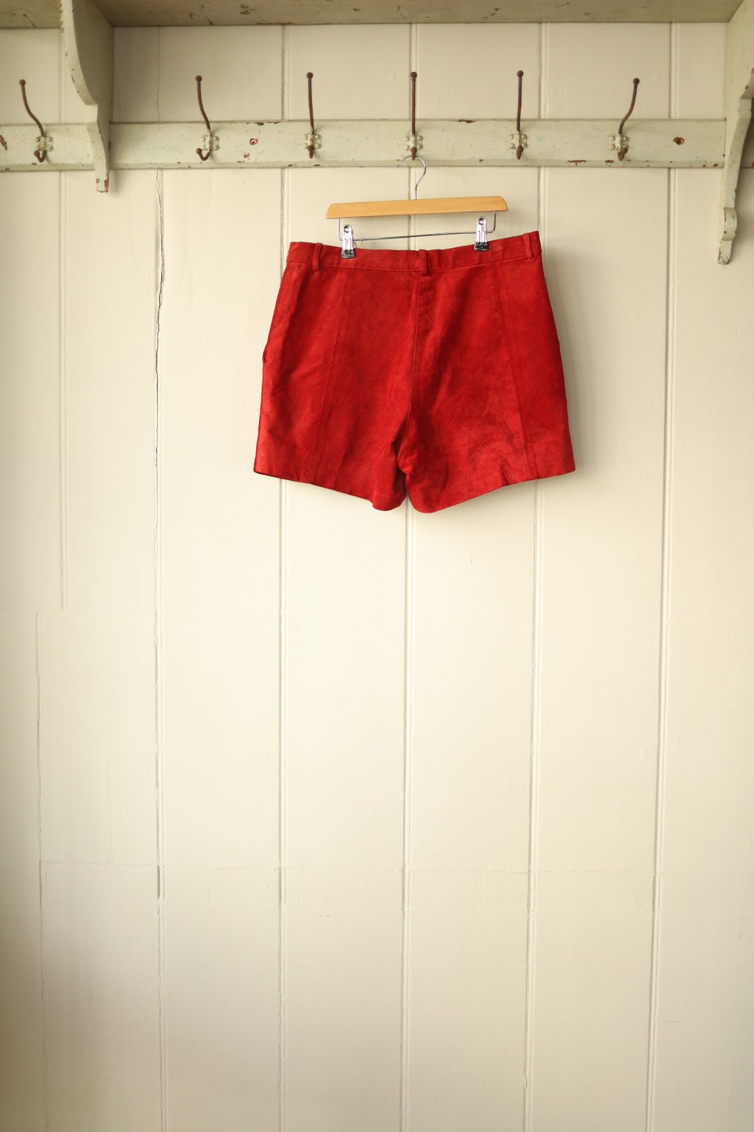 Back view of the red suede shorts showing clean lines and subtle seam detailing, displayed on hanger against cream panelled wall.