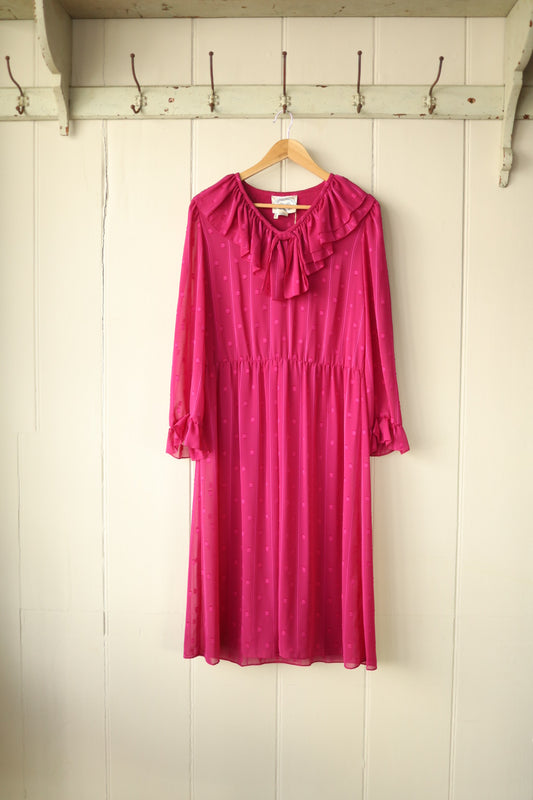 Front view of a raspberry pink sheer dress with polka dot texture, frill collar and gathered waist, hanging on a wooden hanger against a cream panelled wall.
