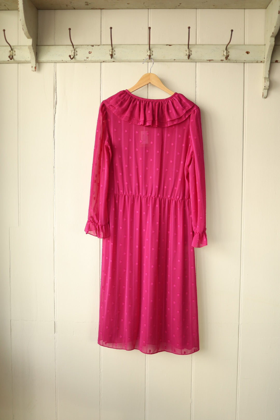 Back view of the raspberry pink sheer dress showing gathered waist and frill collar detail, displayed against a cream panelled wall.