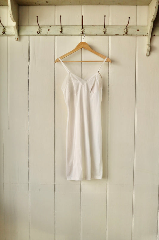 Front view of a vintage cloud-coloured nylon slip with adjustable straps and soft darted bust, hanging on a wooden hanger against a cream panelled wall.